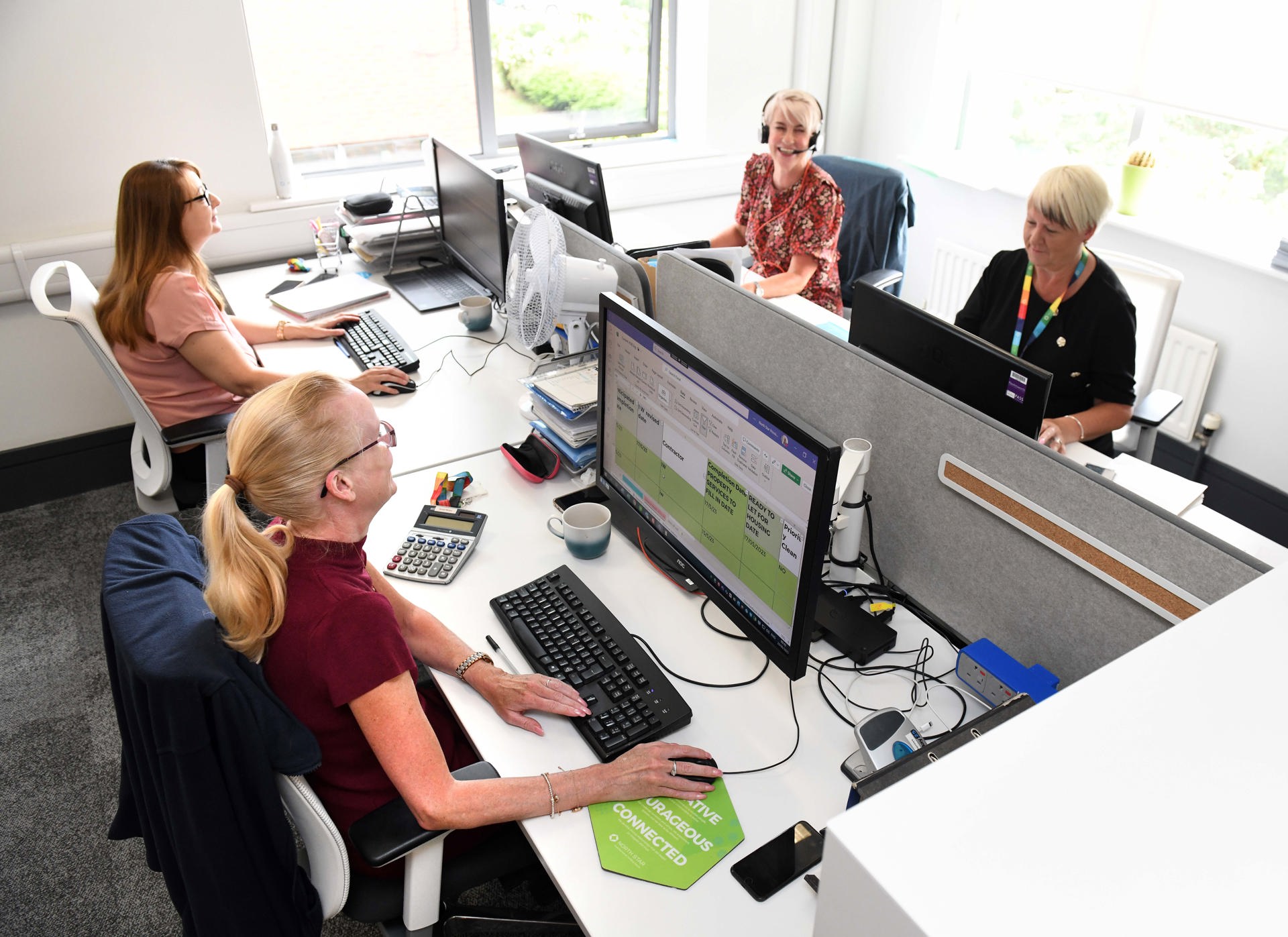 Four women are working at their desks in a bright, modern office. They are using desktop computers and appear focused and engaged. One woman is wearing a headset, and others are smiling and interacting. Office supplies, mugs, and documents are neatly arranged on the desks. A green sign on one desk displays the words "Creative, Courageous, Connected," reflecting North Star's values.