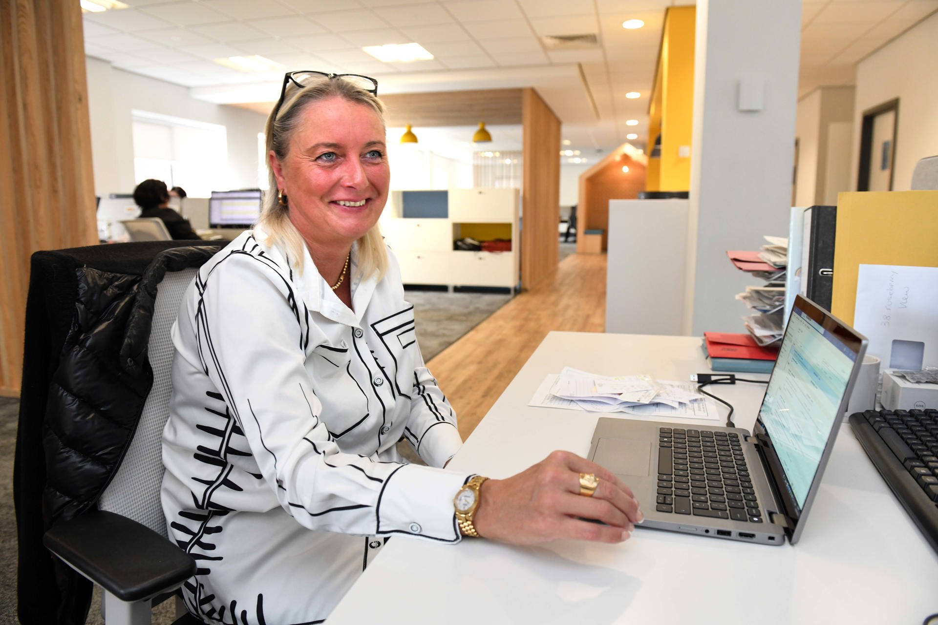  A smiling woman sits at a desk in a bright, modern office space, working on a laptop. She is wearing a white blouse with black patterns and has a pair of glasses resting on her head. Papers and folders are neatly stacked beside her, and the office features a clean, open layout with warm wooden accents