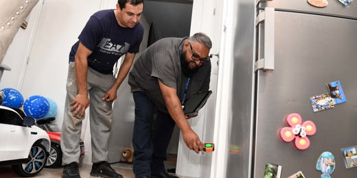 A housing officer uses a handheld device to inspect a wall inside a home, while the resident watches closely. They are in a kitchen area with a fridge covered in magnets and children’s toys visible in the background.  Let me know if you want a version tailored for accessibility compliance or a shorter version for web use.