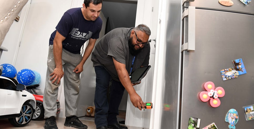 A housing officer uses a handheld device to inspect a wall inside a home, while the resident watches closely. They are in a kitchen area with a fridge covered in magnets and children’s toys visible in the background.  Let me know if you want a version tailored for accessibility compliance or a shorter version for web use.