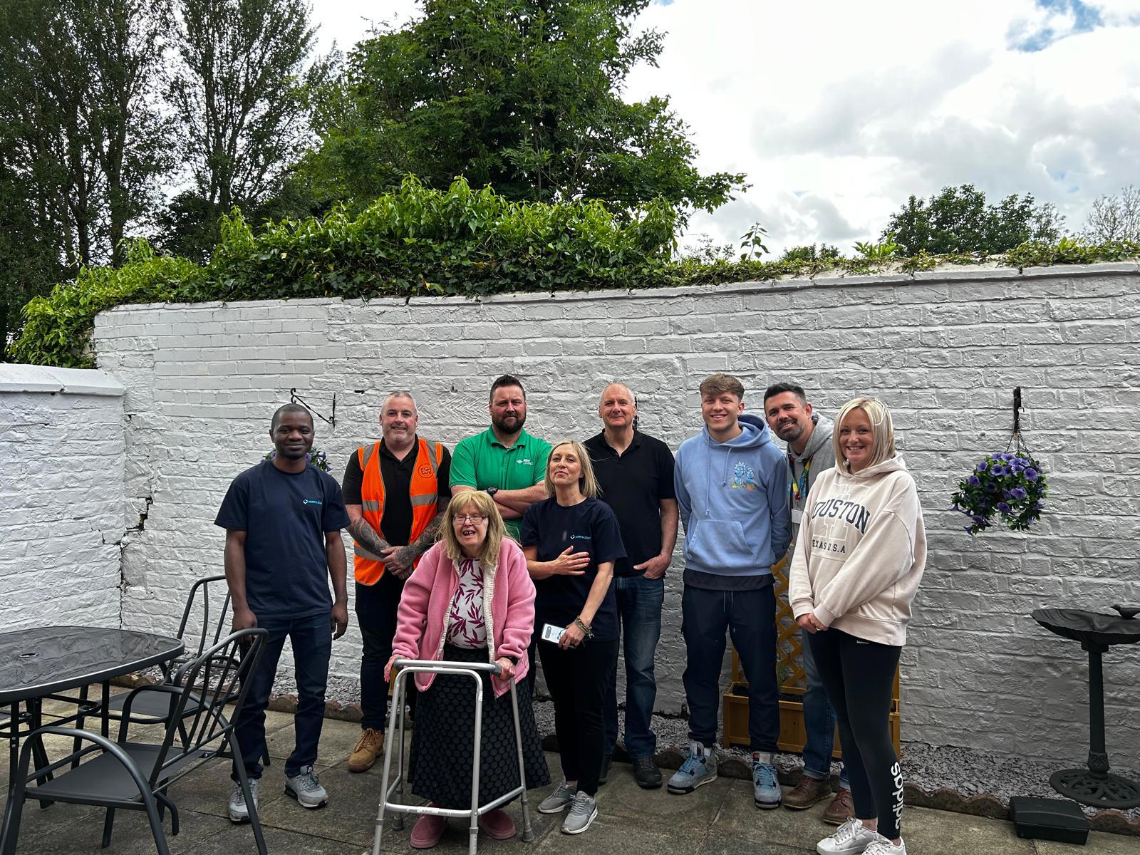A group of ten people posing outdoors in front of a white brick wall and greenery. The group includes a mix of men and women, some in casual clothing and others in work uniforms or high-visibility vests. A woman using a walking frame is at the front center, smiling alongside the group. The setting appears to be a garden or patio area with outdoor furniture and hanging flowers visible