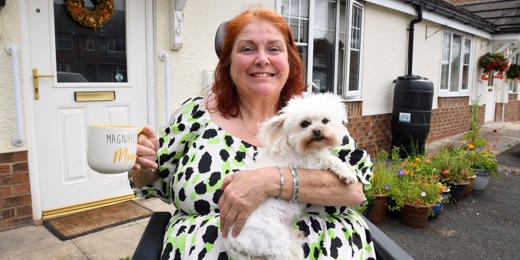 A smiling woman sitting in a wheelchair outside her home, holding a fluffy white dog on her lap and a large mug that reads 'Magnificent Mum.' She is wearing a white dress with green and black spots. Behind her is a white front door with a floral wreath and a garden with colorful potted flowers.