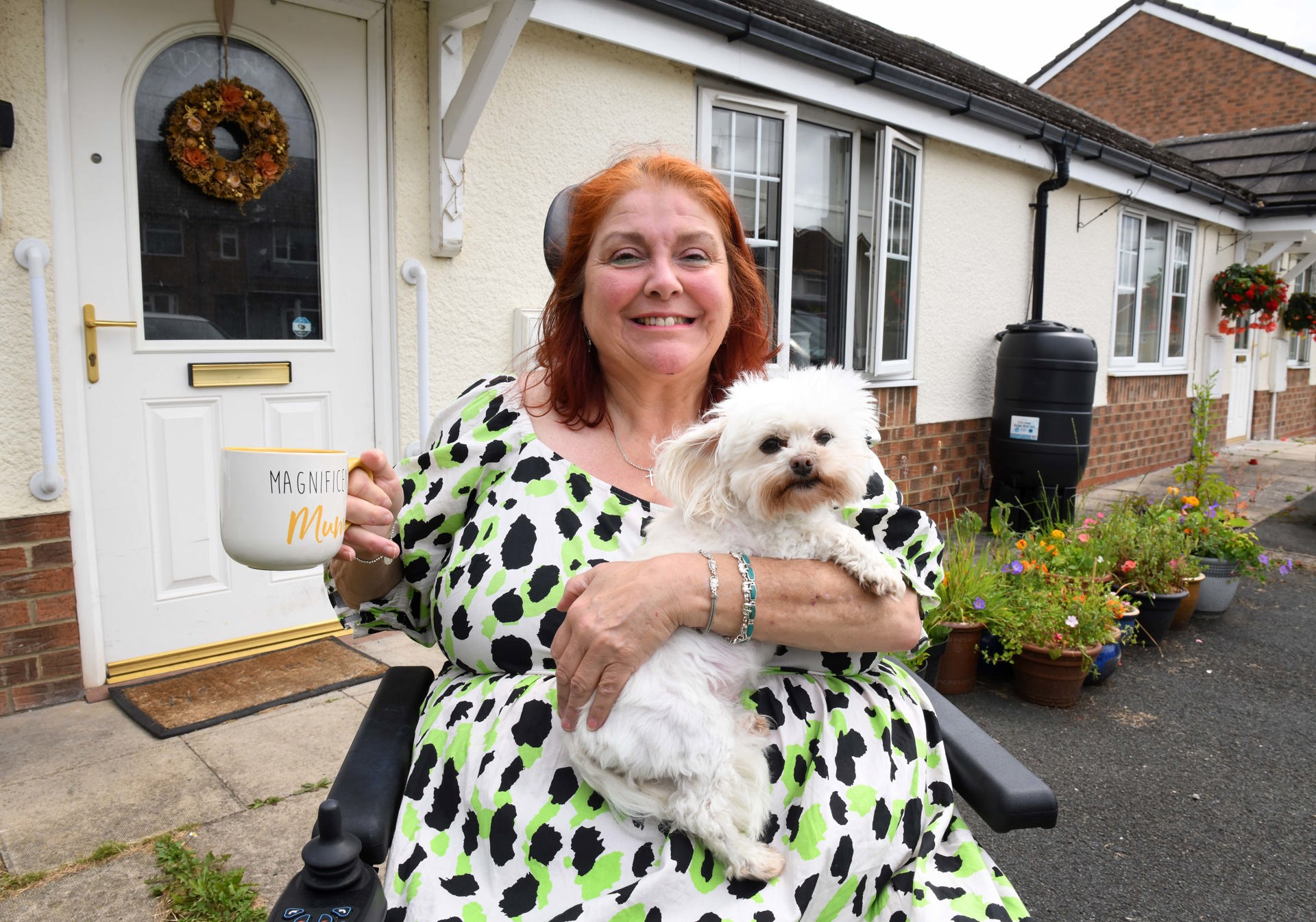 A smiling woman sitting in a wheelchair outside her home, holding a fluffy white dog on her lap and a large mug that reads 'Magnificent Mum.' She is wearing a white dress with green and black spots. Behind her is a white front door with a floral wreath and a garden with colorful potted flowers.